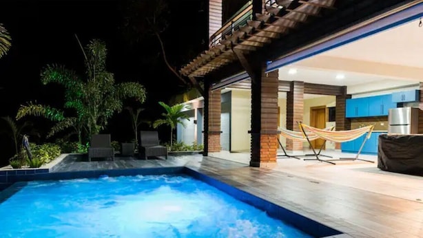 Una piscina con una hamaca y zona de estar exterior, rodeada de plantas y el interior de una casa moderna.