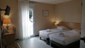 A hotel room with two beds, a desk with a lamp, a chair, and a view of the outside. - Hôtel du Commerce (Tronget)