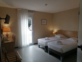 Quarto de hotel com duas camas, uma escrivaninha com abajur, uma cadeira e vista para o exterior.