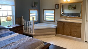 Ein Schlafzimmer mit einem großen Bett, einer hölzernen Bank, einem Fenster mit Jalousien, einem kleinen Tisch mit Lampe sowie einem Badezimmer mit Badewanne, Waschbecken und Holzschränken.