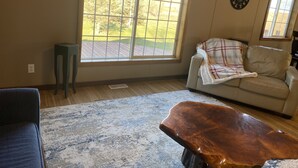 Ein Wohnzimmer mit großem Fenster, einem Holztisch, einer Couch mit Decke und einer Uhr an der Wand.