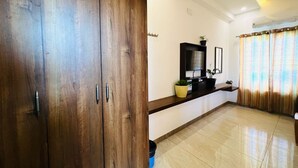 A spacious room with a wooden wardrobe, a flat-screen TV, a small shelf with plants, and a large window with curtains. - Raja residency near ISHA ADIYOGI ALANDURAI Siruvani Coimbatore ,Tamilnadu, India (Alanthurai)