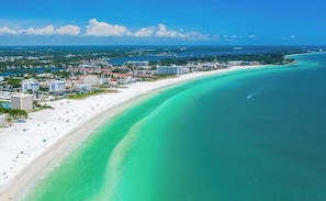 An aerial view of a coastal area with a beach, buildings, and clear turquoise waters.