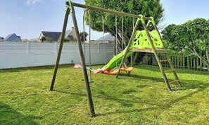 A swing set with a slide, a ladder, and a pink ball in a grassy yard with a white fence and trees in the background.