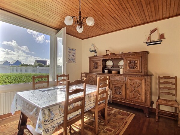 A dining area with a table set for four, wooden chairs, a patterned tablecloth, a wooden cabinet with decorative elements, and a view of the sea through a large window. - Cottage d'Ys in Pentrez (500 m from the beach) (Saint-Nic)