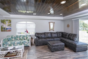 Uma sala de estar com sofá em formato de L, uma mesinha com cadeiras, um espelho decorativo e uma janela com persianas.