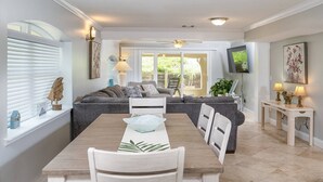 Ein Essbereich mit Holztisch, weißen Stühlen und Blick auf ein Wohnzimmer mit Sofa und Fernseher.