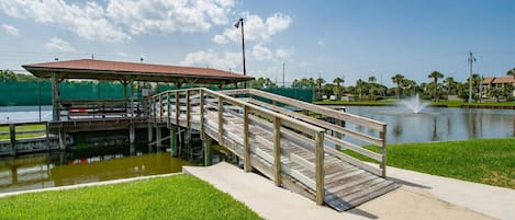 A wooden bridge leading to a covered pavilion over a body of water, with a fountain in the background.