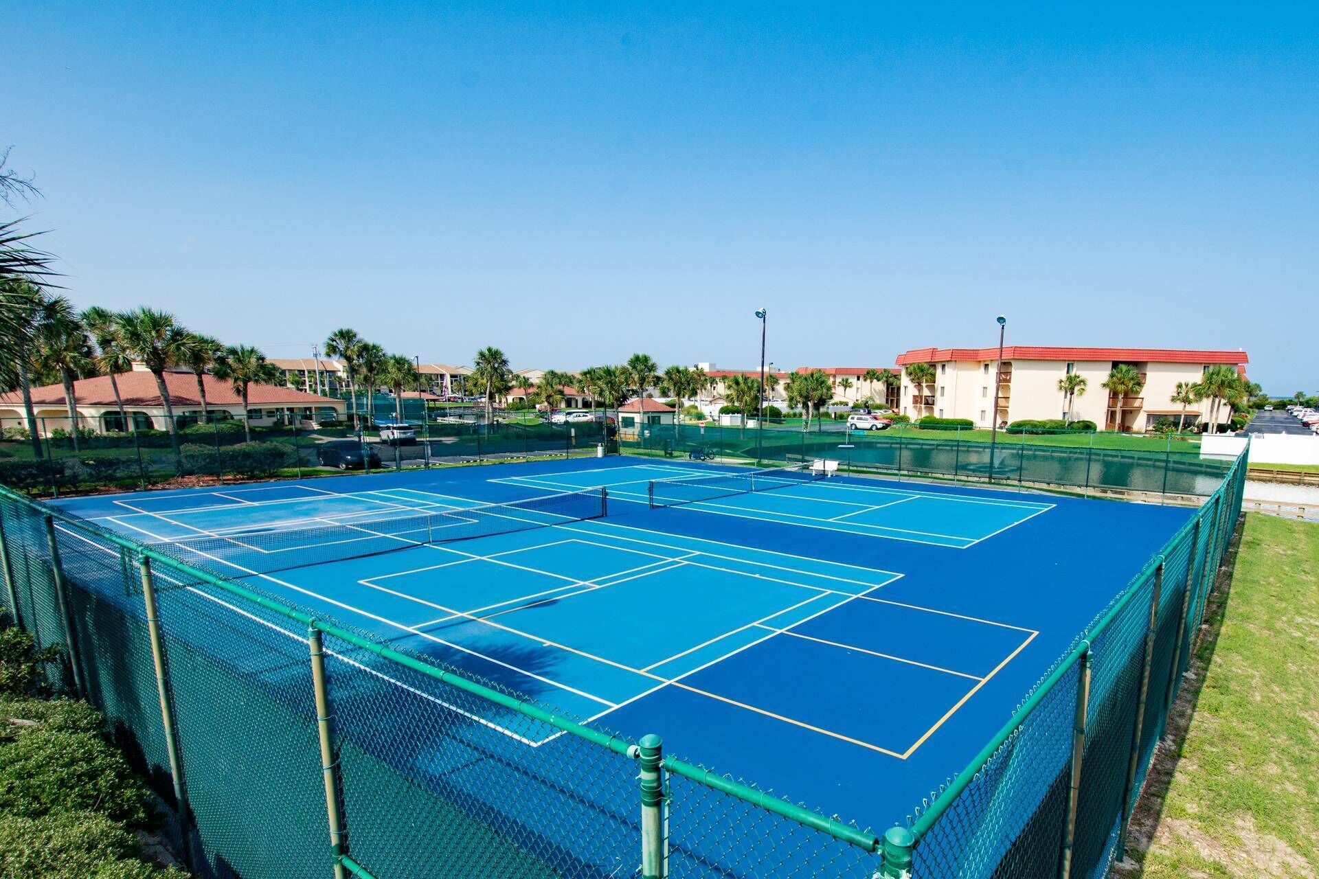 A tennis court with a net, surrounded by a green fence, and adjacent to a body of water.