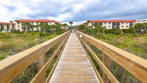 A wooden boardwalk leading to a row of buildings with red roofs, surrounded by greenery. - Ocean and Racquet 8208 (St. Augustine)