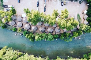 An aerial view of a riverside resort with thatched-roof huts, a parking area, and a clear river.
