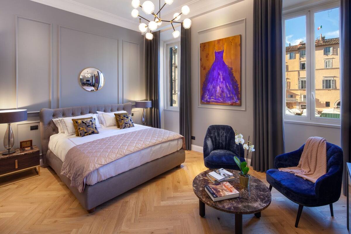A modern bedroom with a large bed, a round mirror, a painting, a chair, and a view of buildings through a window.