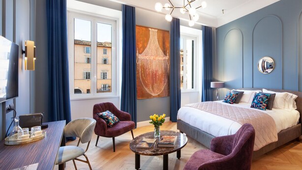 Una habitación de hotel moderna con una cama grande, un escritorio con una computadora portátil, una silla, una mesita de centro con un jarrón de flores y un ventanal con cortinas.