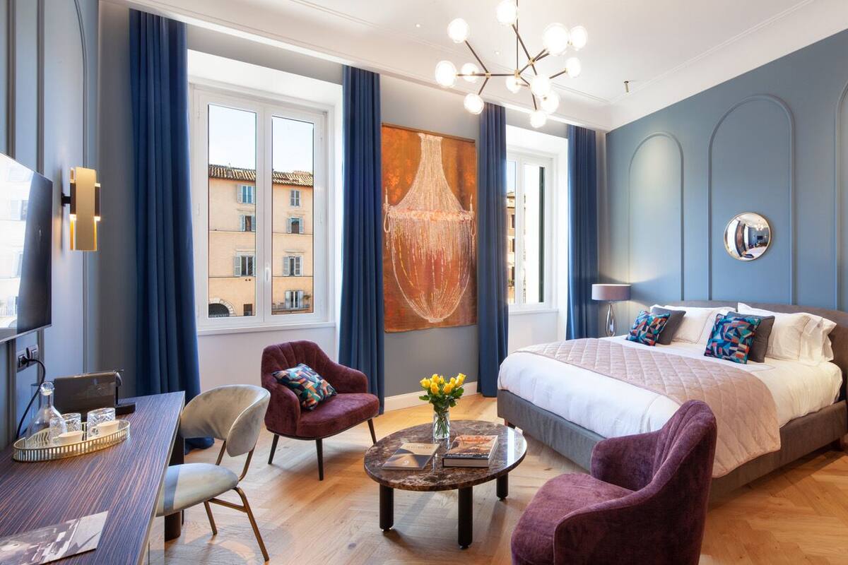 A modern hotel room with a large bed, a desk with a laptop, a chair, a coffee table with a vase of flowers, and a large window with curtains.