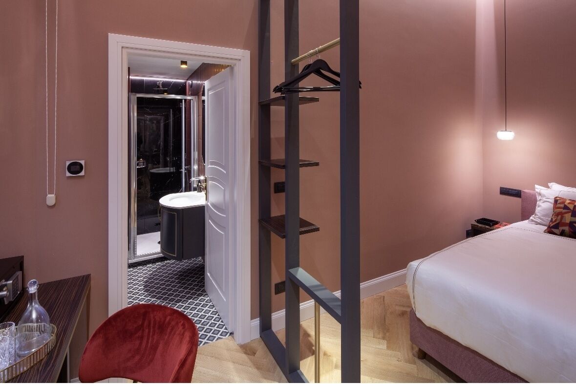 A hotel room with a bed, a chair, a small table with glasses, a hanging coat rack, and a bathroom with a bathtub and sink.