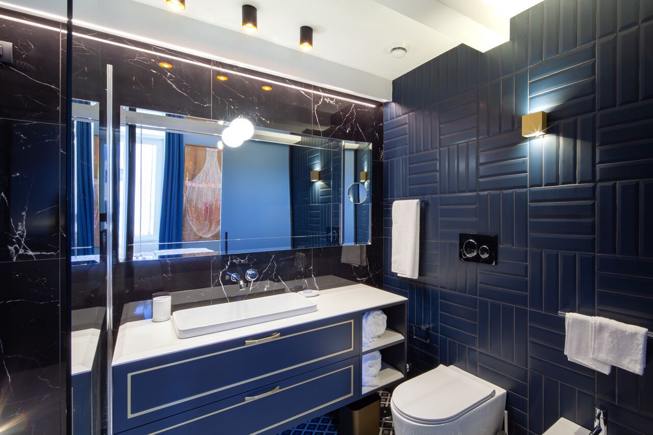 Un baño moderno con un revestimiento de azulejos azul oscuro, un lavamanos blanco con encimera de mármol, un espejo grande y un inodoro.