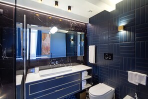 A modern bathroom with a dark blue tiled wall, a white sink with a marble countertop, a large mirror, and a toilet. - Migliori Collection Piazza Navona 71 (Rome)