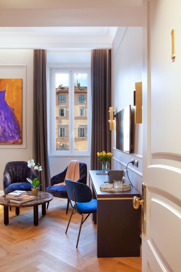 A modern hotel room with a wooden herringbone floor, a large window with curtains, a flat-screen TV, a desk with a vase of flowers, and two chairs. - Migliori Collection Piazza Navona 71 (Rome)