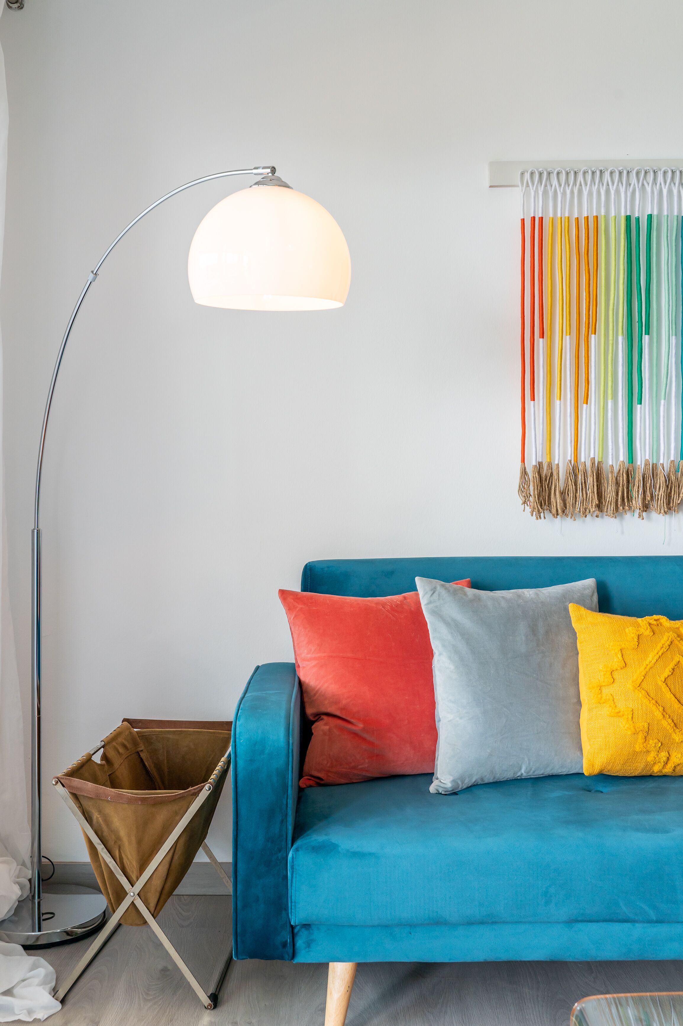 A modern living room with a blue sofa, colorful cushions, a floor lamp, and a decorative wall hanging.