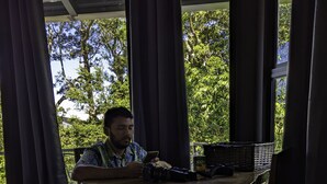 A person sitting at a wooden table with a laptop, under a covered area with curtains, with trees visible outside. - Quality Cabins Monteverde #4 (Monteverde)