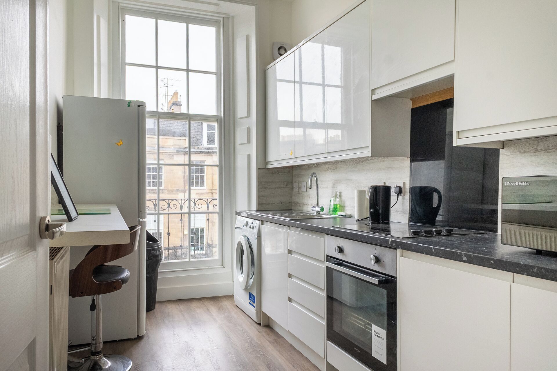 A compact kitchen with white cabinets, a built-in oven, a washing machine, and a window overlooking a cityscape.