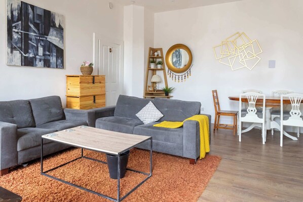 A living room with a grey sofa, a wooden coffee table, and a dining area with a wooden table and white chairs.
