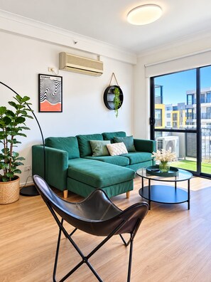 A modern living room with a green sectional sofa, a black leather chair, a glass coffee table, and a large window offering a view of buildings.