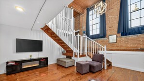 A modern living room with a wooden staircase, a flat-screen TV, a fireplace, and a brick wall. - Stylish Loft with Modern Amenities in Detroit (Detroit)
