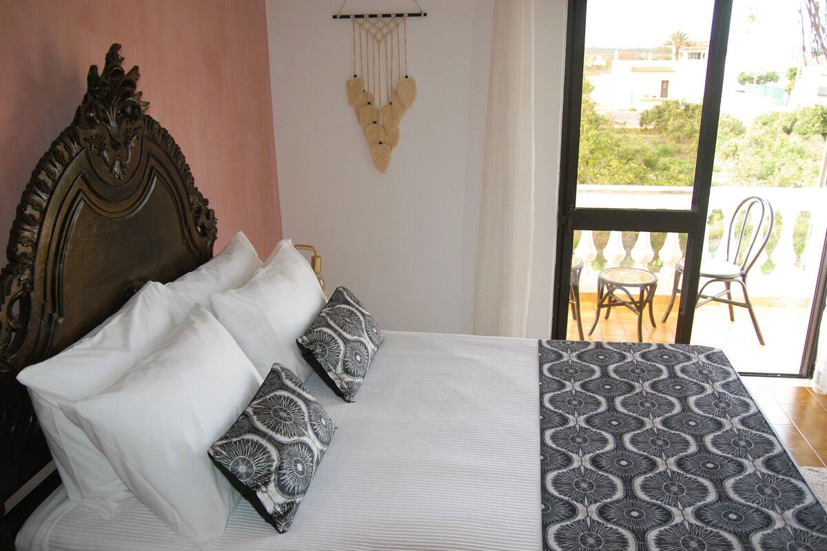 A bed with white and patterned pillows, a decorative wall hanging, and a view of a balcony with chairs.