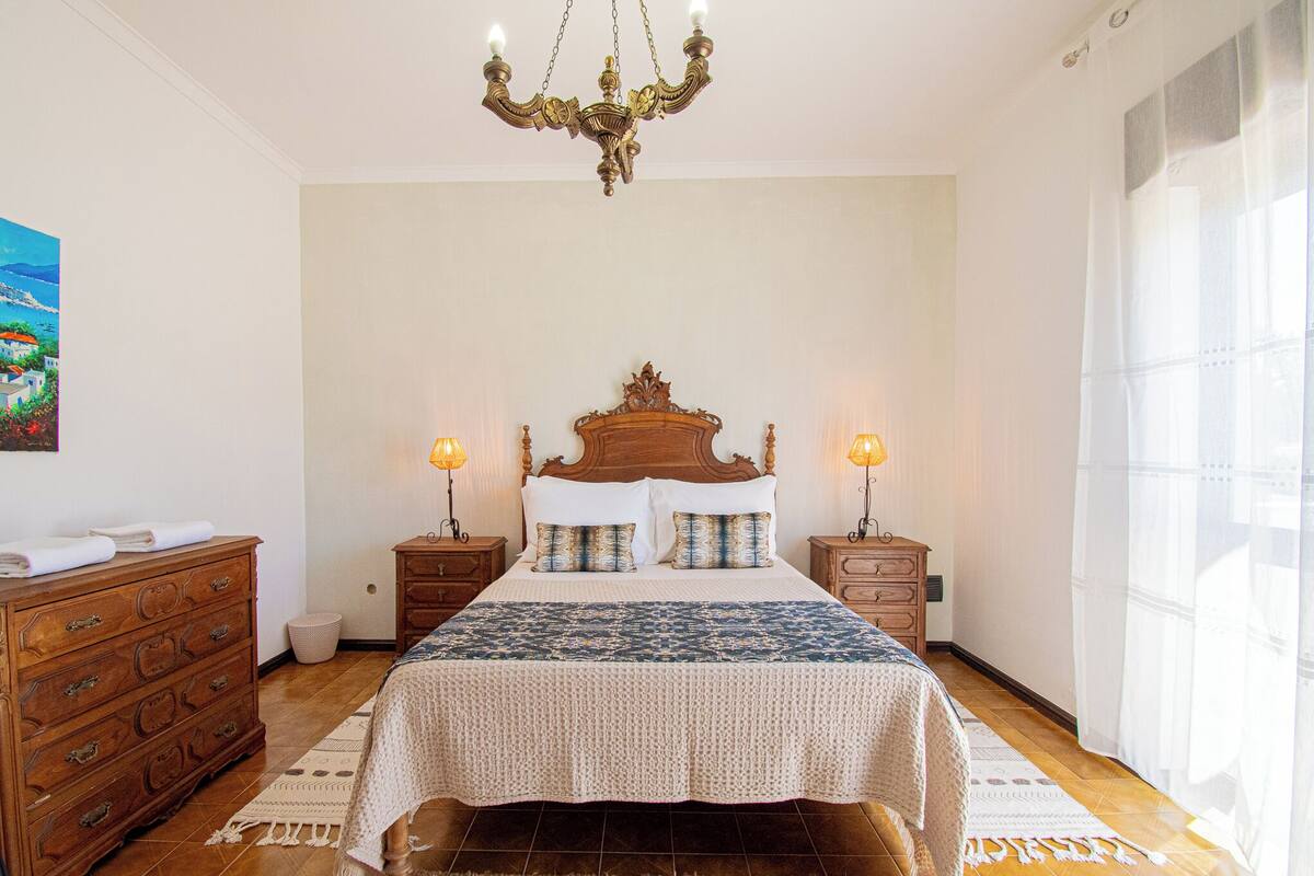 A bedroom with a large bed, wooden headboard, two bedside tables with lamps, a dresser, and a painting on the wall.