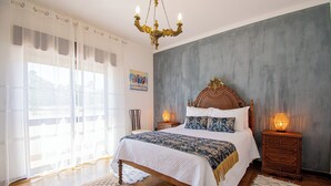 A bedroom with a large bed, a chandelier, a nightstand with a lamp, and a window with sheer curtains.