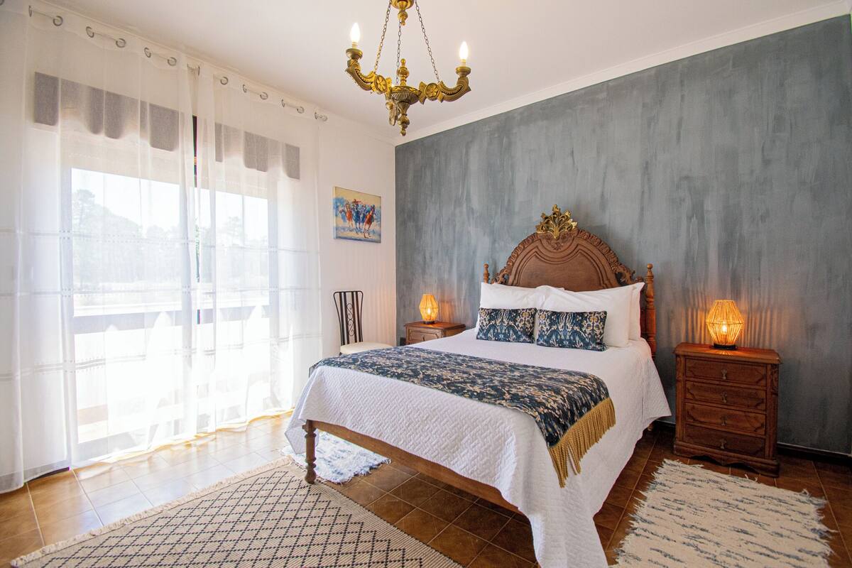 A bedroom with a large bed, a chandelier, a nightstand with a lamp, and a window with sheer curtains.