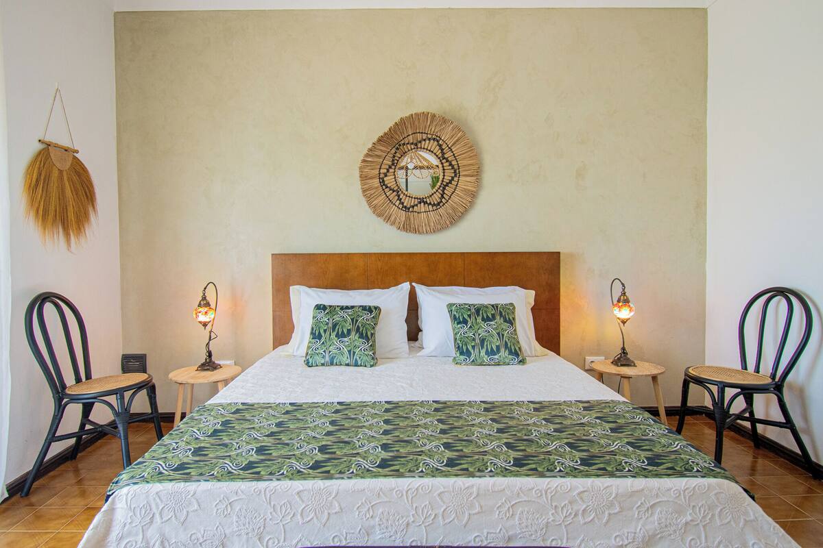 A neatly made bed with a patterned bedspread, two matching pillows, and a round wall hanging above. There are two small tables with lamps beside the bed, and two wicker chairs.