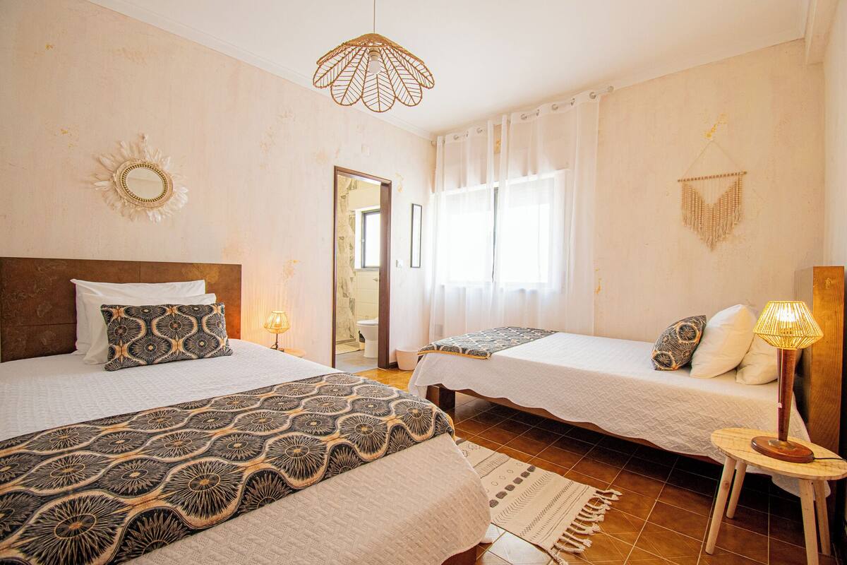 A hotel room with two beds, a large mirror, a hanging light fixture, and a window with curtains.