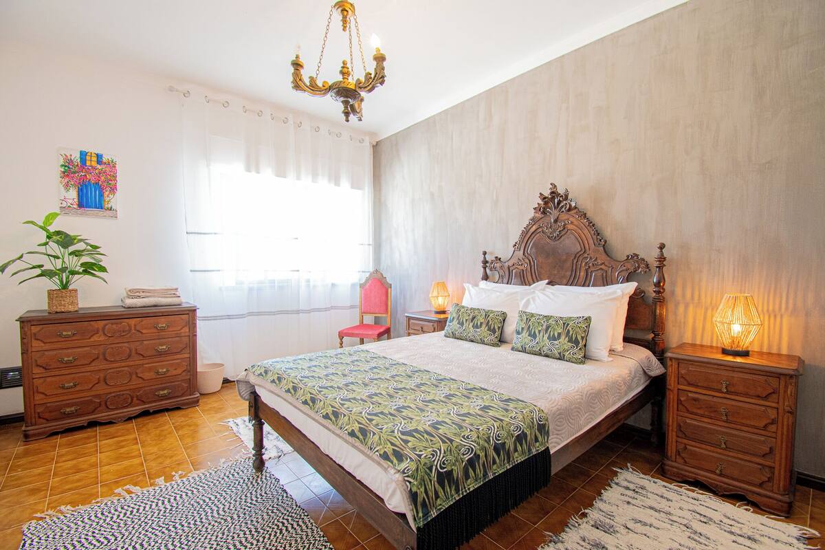 A bedroom with a large bed, wooden nightstands, a chandelier, a potted plant, and a window with curtains.