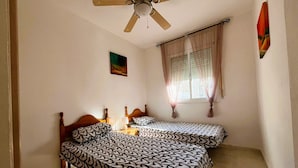 A room with two beds, a ceiling fan, a window with curtains, and a small painting on the wall. - Villa Jane (Les Tres Cales)
