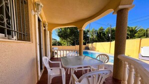 A covered outdoor seating area with a pool, white plastic chairs, and a glass table. - Villa Jane (Les Tres Cales)