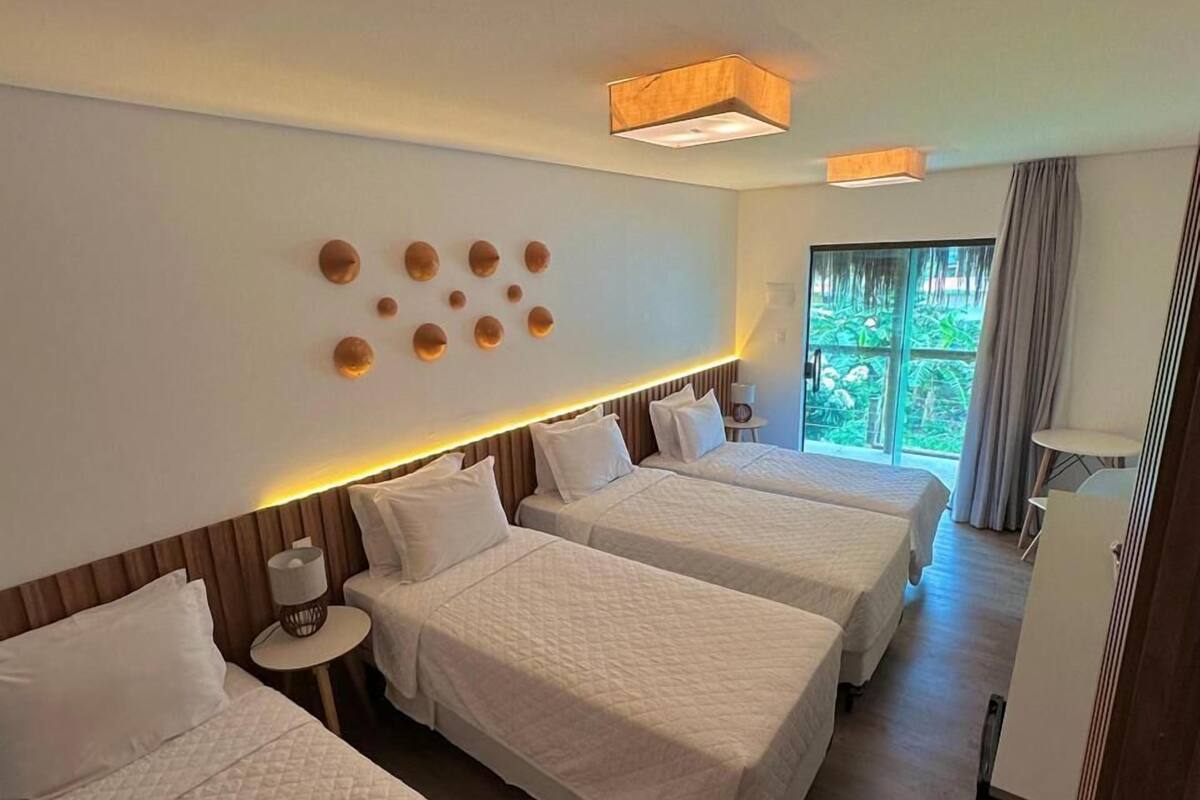 A hotel room with three beds, a large window, and a view of greenery outside.