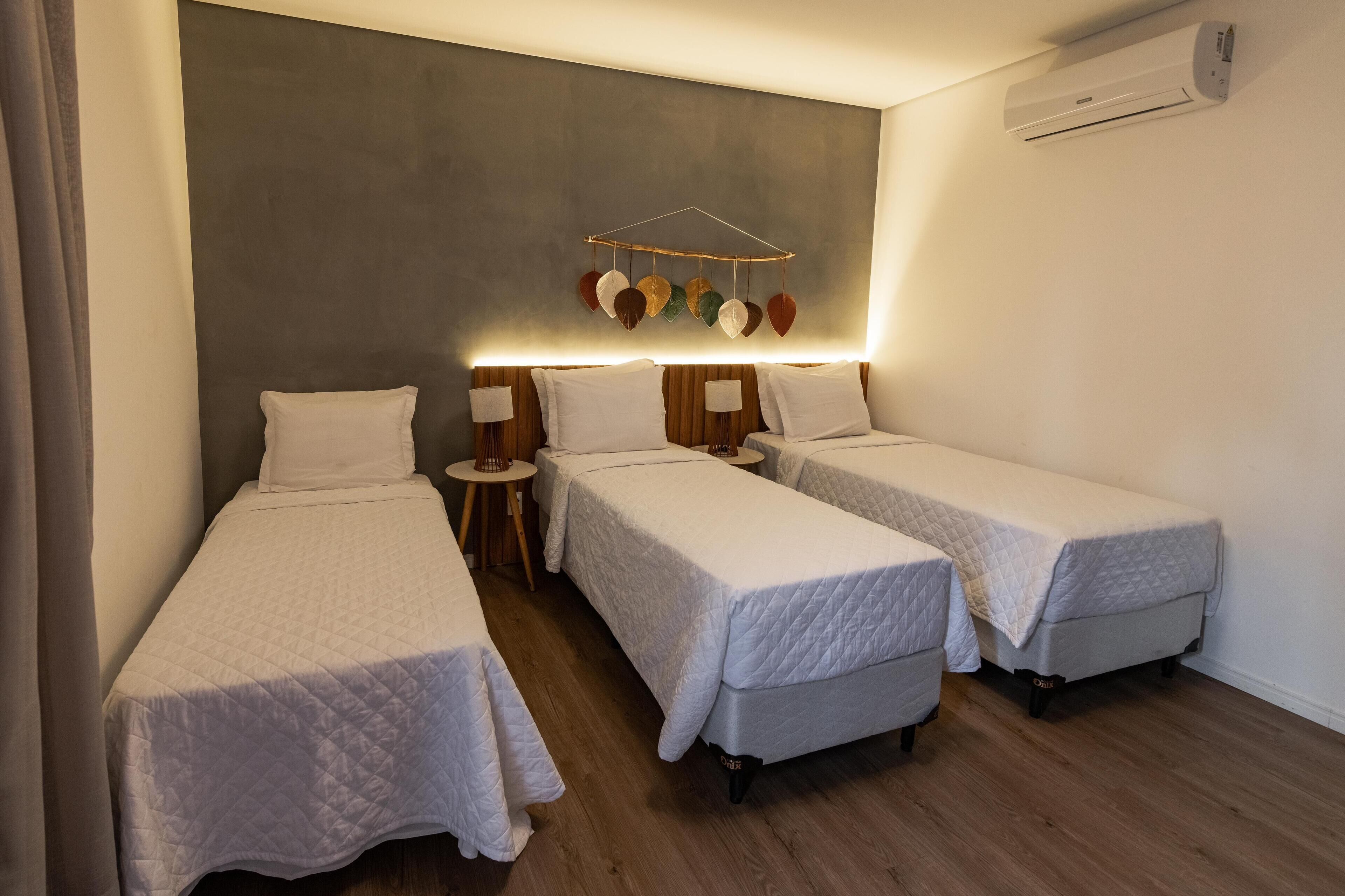 Ein Hotelzimmer mit drei Einzelbetten, einem Nachttisch mit Lampe, einer Wandklimaanlage und einem dekorativen Wandbehang.