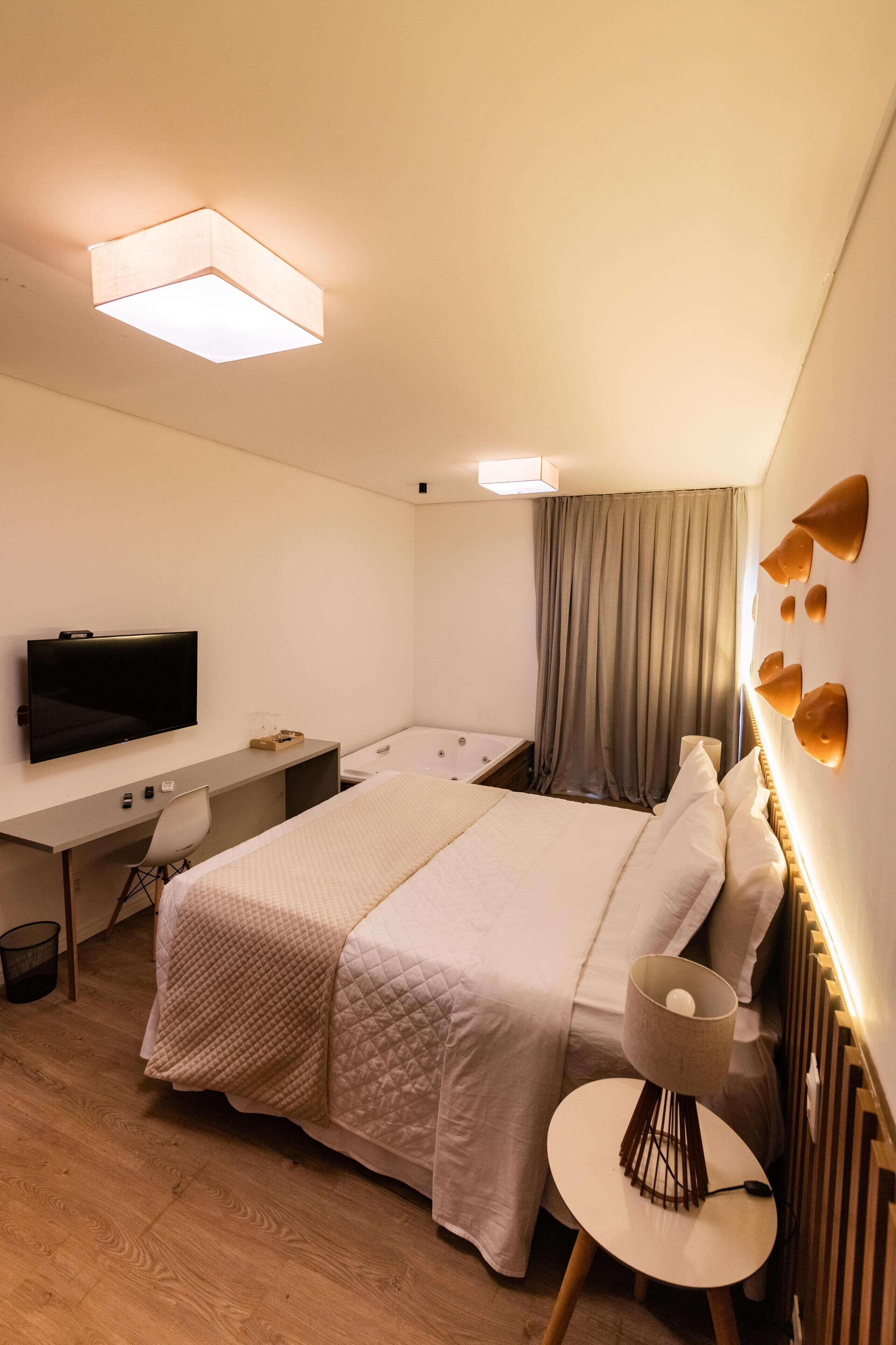 Habitación de hotel con cama, escritorio, televisor, silla, lámpara y ventana con cortinas.