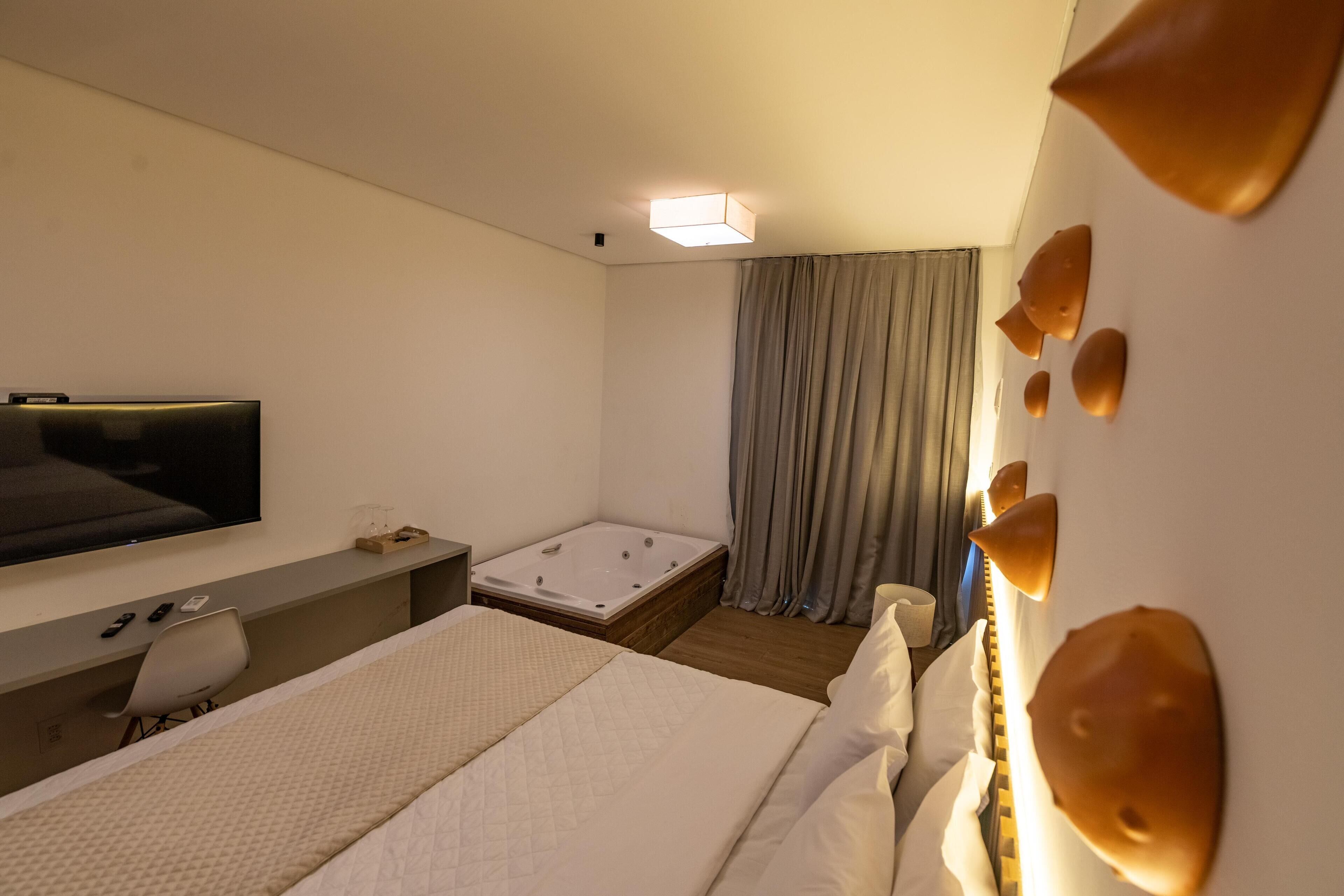 Habitación de hotel con cama, escritorio, televisión, baño y un ventanal con cortinas.