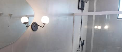 Moderni kylpyhuone, jossa on suuri peili, valkoinen pesuallas, wc ja lasiseinällä erotettu suihkutila.