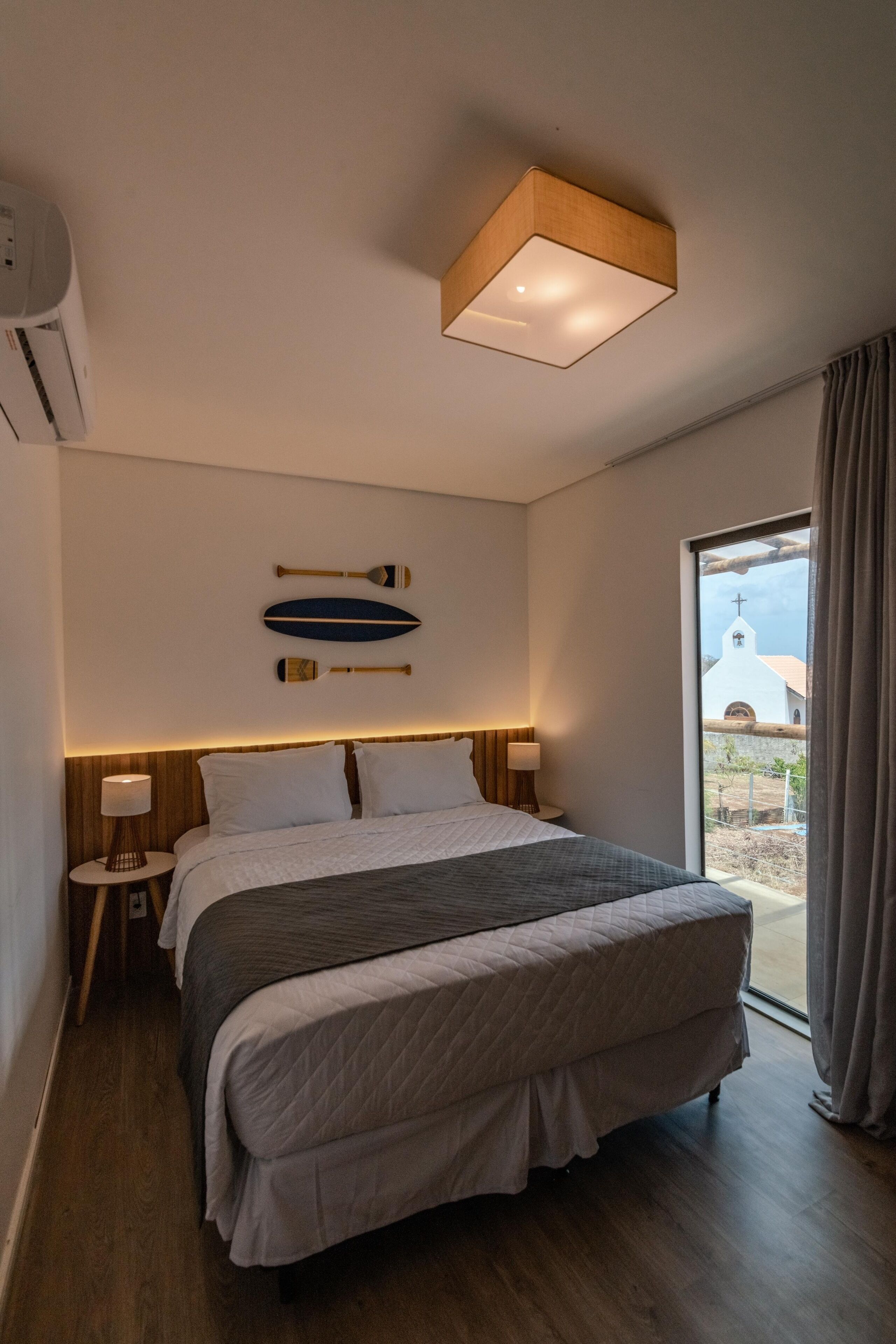 A modern hotel room with a large bed, a wooden headboard, a wall-mounted light fixture, and a view of a church through the window.