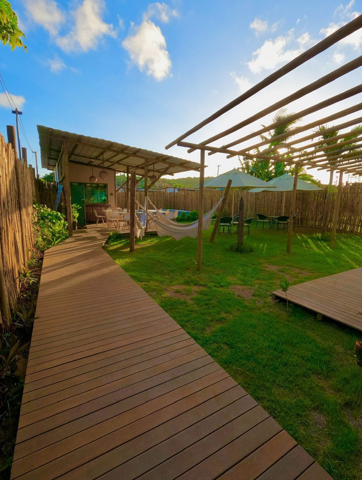 A wooden deck leading to a covered outdoor area with a hammock and tables, surrounded by a bamboo fence and greenery.