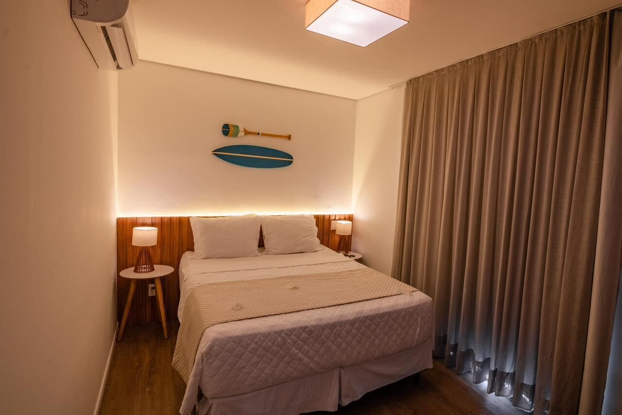 A hotel room with a bed, wooden headboard, two bedside lamps, a wall-mounted decorative item, and a window with curtains.