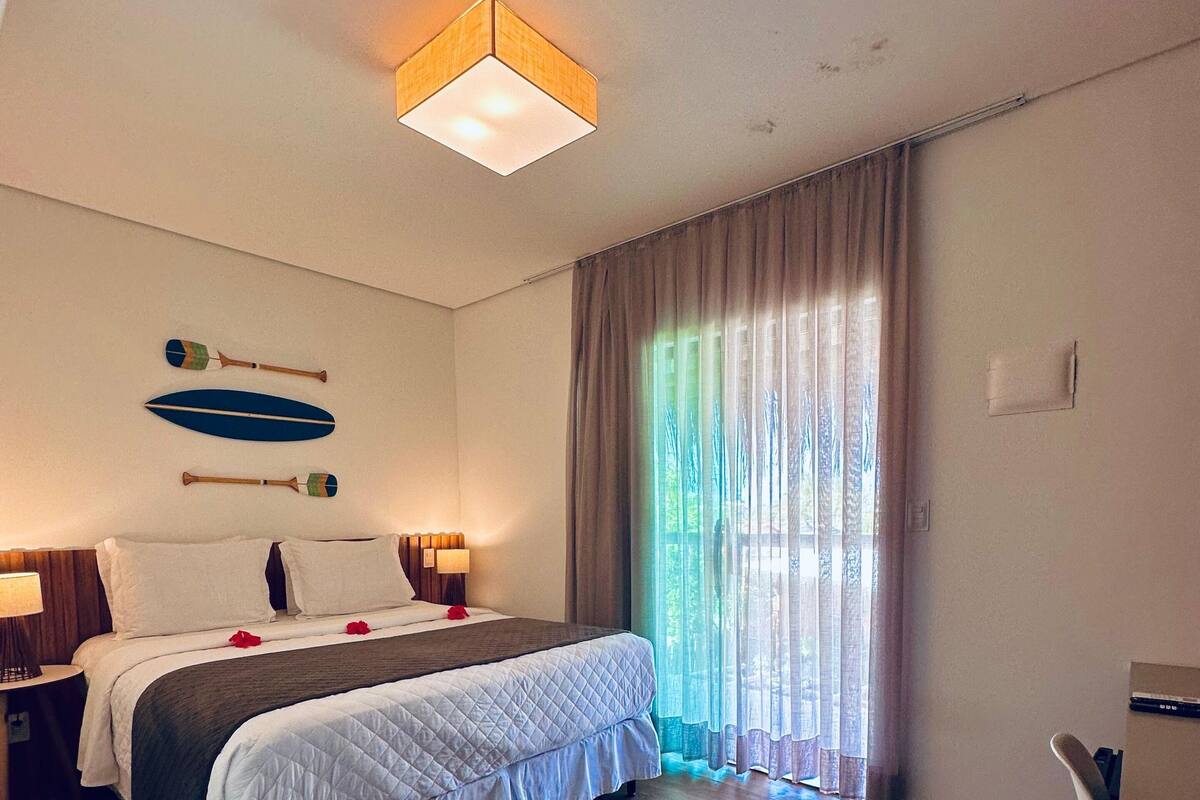 A hotel room with a large bed, a bedside table, a lamp, a ceiling light, and a window with curtains.