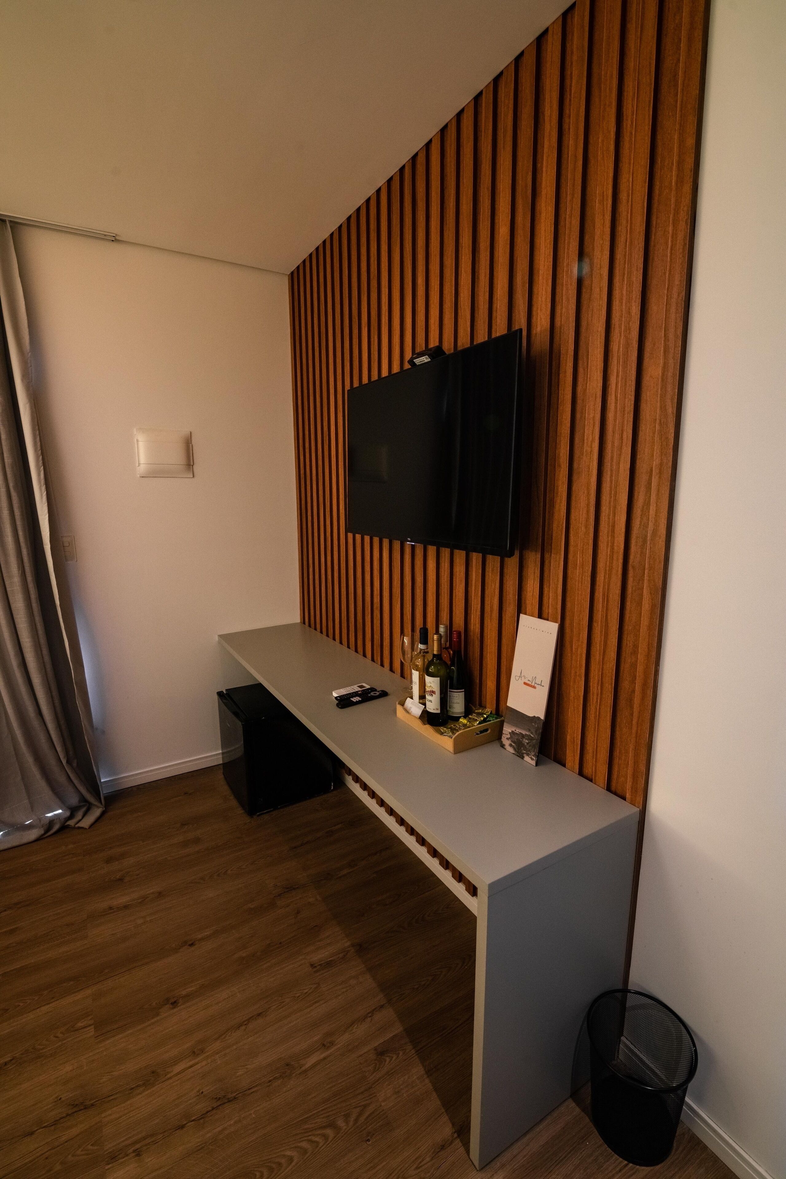 A modern room with a wooden slat wall, a mounted TV, a small desk with a trash bin, and a window with curtains.