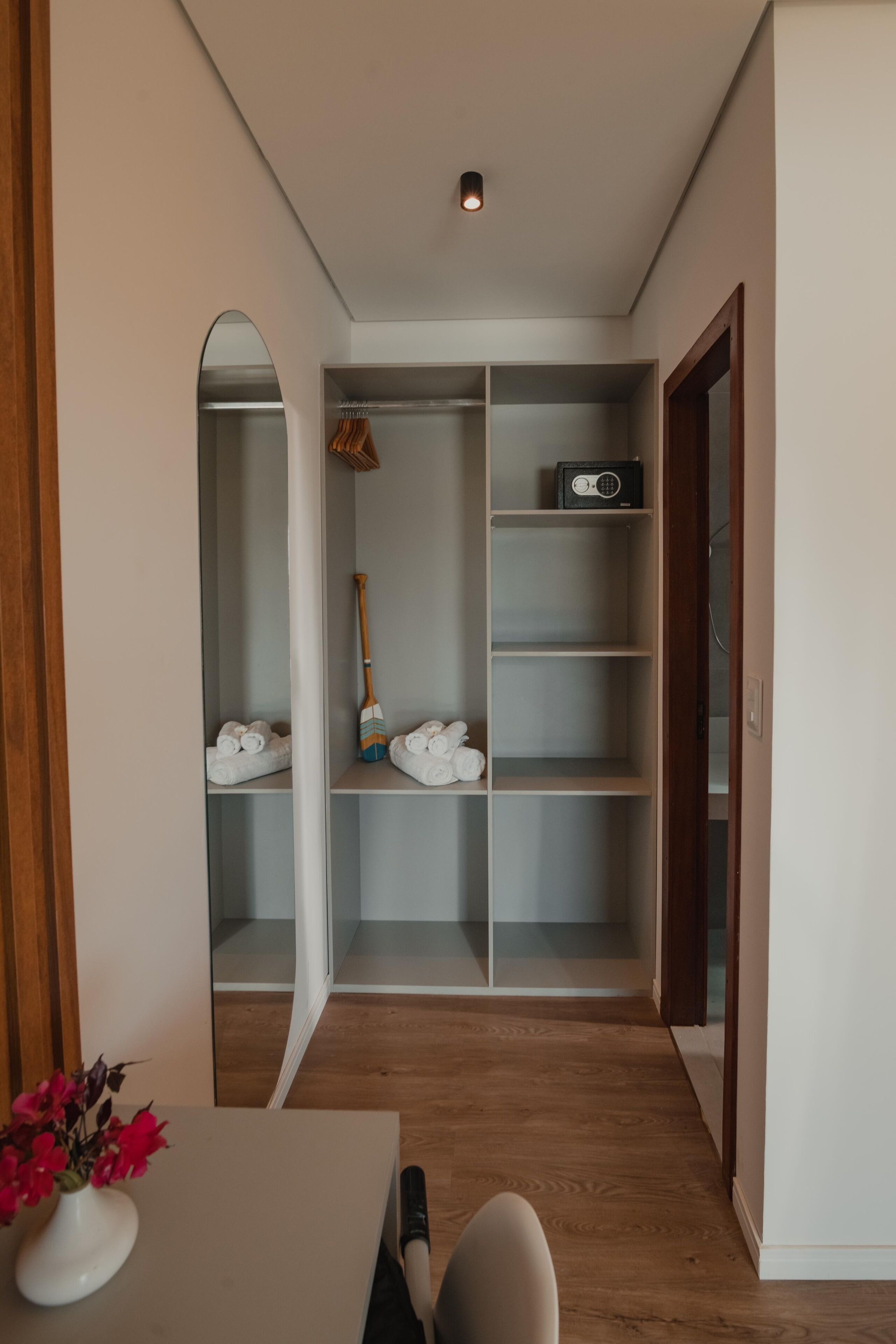 A hallway with a wooden floor, a white table with a vase of flowers, and a closet with shelves and a hanging rack.
