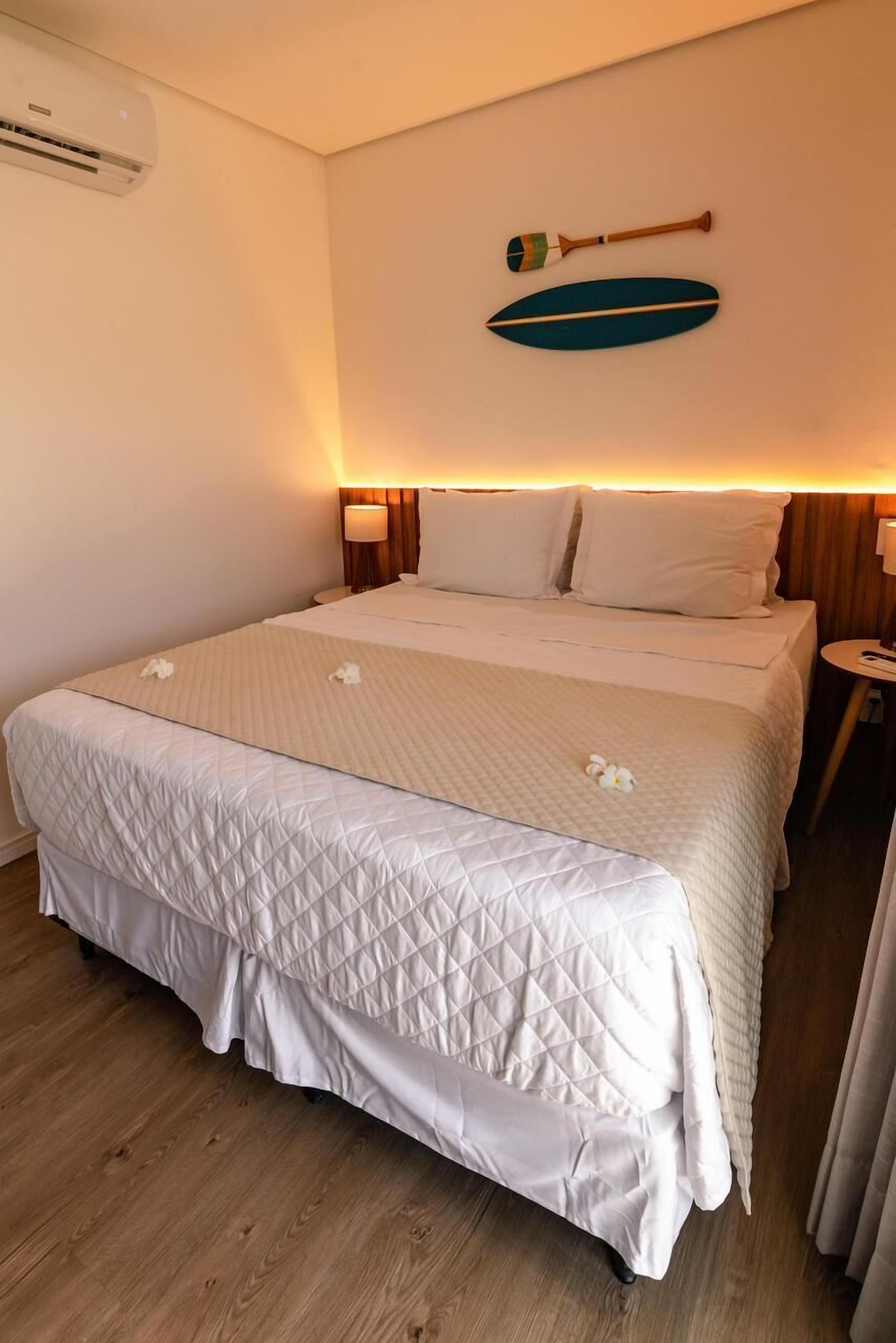 A hotel room with a large bed, a wall-mounted air conditioner, and a wooden nightstand with a lamp.
