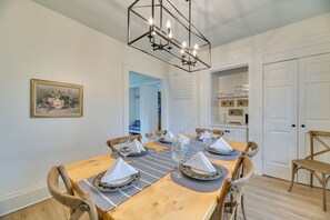 Comedor con una mesa de madera dispuesta para seis comensales, rodeada de sillas, una araña y un cuadro con flores en la pared.